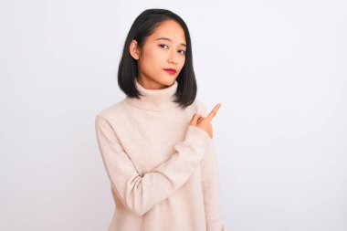 Balıkçı yaka kazak giyen genç bir kadın izole edilmiş beyaz arka planda duruyor. El parmağı ile yan tarafı işaret ediyor. Ciddi ve sakin bir yüz ifadesi var.