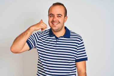Sıradan çizgili polo giyen genç bir adam izole edilmiş beyaz arka planda durup telefonla konuşurken gülümseyerek el ve parmaklarıyla telefon hareketi yapıyor. İletişim kavramları.