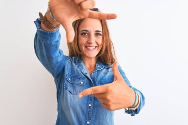 İzole edilmiş arka planda duran kot ceketli genç kızıl saçlı kadın gülümsüyor ve mutlu yüzlü elleri ve parmakları olan bir çerçeve yapıyor. Yaratıcılık ve fotoğrafçılık kavramı.