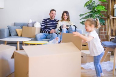 Güzel aile, anne-babalar kanepede kahve içme oturmuş karton kutular etrafında yeni evde oynayan çocuğuna bakıyor