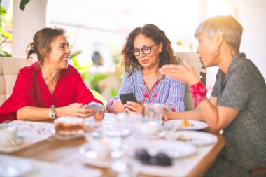 Orta yaşlı kadınlarla buluşup, öğle yemeği yiyip, kahve içiyorlar. Olgun arkadaşlar mutlu bir şekilde gülümsüyor. Güneşli bir günde evde akıllı telefon kullanıyorlar.