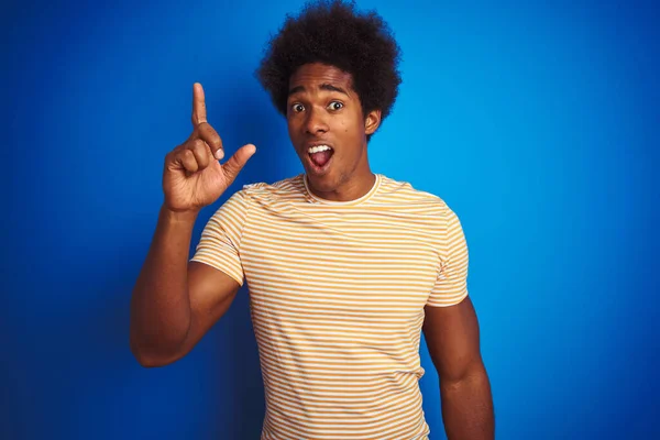 アフロの髪をしたアメリカ人男性は 青い背景の上に縞模様の黄色のTシャツを着て 成功したアイデアで指を指しています 興奮し 第一番 — ストック写真