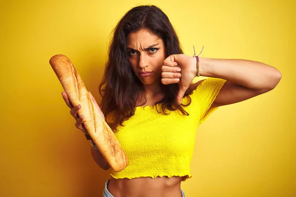 パンを持っている若い美しいパン屋の女性は怒っている顔で孤立した黄色の背景の上に立っています 親指で嫌いを示す負の兆候ダウン 拒絶概念 — ストック写真