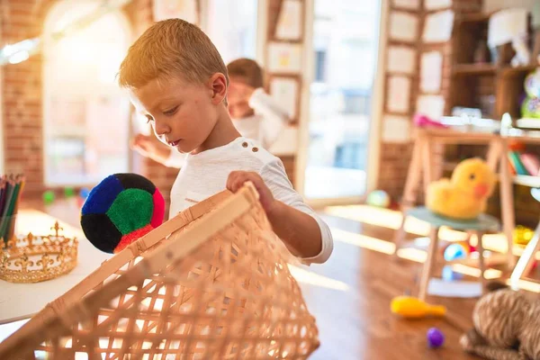 Adorable blonde twins playing basketball using wicker basket and ball around lots of toys at kindergarten