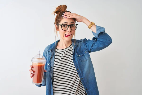 Beautiful redhead woman drinking smoothie of tomato over isolated white background stressed with hand on head, shocked with shame and surprise face, angry and frustrated. Fear and upset for mistake.