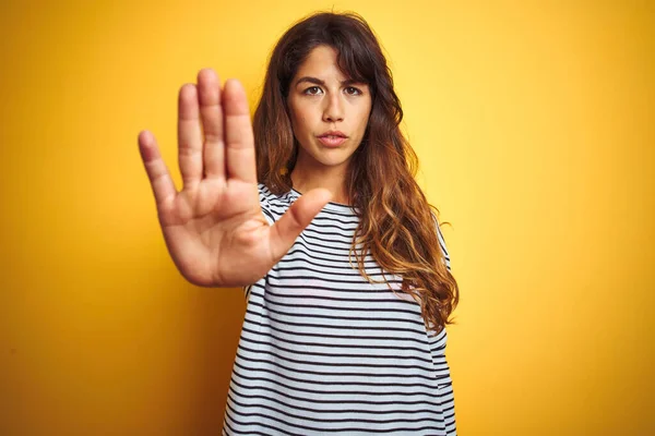 Young beautiful woman wearing stripes t-shirt standing over yelllow isolated background doing stop sing with palm of the hand. Warning expression with negative and serious gesture on the face.