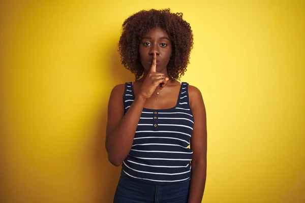 Young african afro woman wearing striped t-shirt over isolated yellow background asking to be quiet with finger on lips. Silence and secret concept.