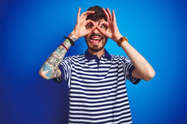 İzole edilmiş mavi arka planda duran çizgili polo giyen genç bir adam dürbün gibi dil çıkartıyor, gözler parmaklarından bakıyor. Çılgın ifade.