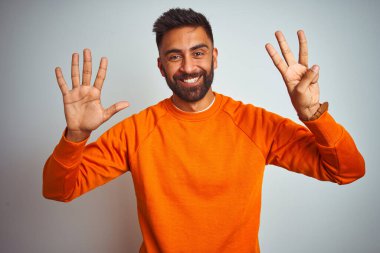 İzole edilmiş beyaz arka planda turuncu süveter giyen genç Hintli adam sekiz numaralı parmaklarıyla gülümseyerek kendinden emin ve mutlu bir şekilde işaret ediyor..