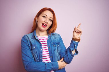 Güzel kızıl saçlı kadın kot gömlek ve çizgili tişört giyerek izole edilmiş pembe arka planda, yüzünde kocaman bir gülümsemeyle kameraya el ile işaret ediyor..