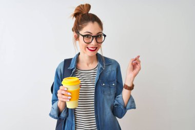 Güzel kızıl saçlı bayan kahve içerken izole edilmiş beyaz arka planda çok mutlu bir şekilde elini ve parmağını kenarı işaret ediyor.