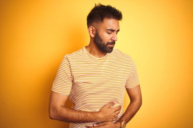 Yalıtılmış sarı arka planda duran tişörtlü genç Hintli adam midesi üstünde duruyor çünkü hazımsızlık, acı veren hastalıklar kendini iyi hissetmiyor. Ache kavramı.