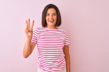 İzole edilmiş pembe arka plan üzerinde duran çizgili tişört giyen genç ve güzel bir kadın üç numaralı parmaklarıyla gülümseyerek kendinden emin ve mutlu bir şekilde onu işaret ediyor..