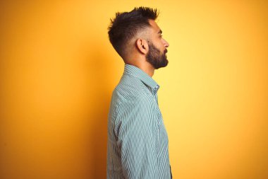 Yeşil çizgili tişört giyen genç Hintli adam, yalıtılmış sarı arka plan üzerinde duruyor, yan yana bakıyor, rahat profil pozu veriyor, kendine güvenen bir gülümsemesi var..