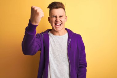 Mor eşofman üstü giyen genç ve yakışıklı bir adam izole edilmiş sarı arka planda kızgın ve kızgın duruyor. Öfkeyle bağırırken yumruğunu kaldırıyor. Öfke ve agresif kavram.