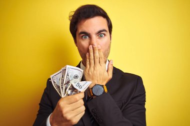Takım elbiseli genç yakışıklı işadamı, dolarları izole edilmiş sarı arka planda tutuyor. Ağzı kapalı, utançtan ağzı açık, korkunun dışa vurumu, sessizlikten korkmak, gizli konsept.