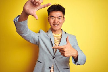 Asyalı genç iş adamı ceketini giyip izole edilmiş sarı arka planda duruyor gülümsüyor ve mutlu yüzlü elleri ve parmakları olan bir çerçeve yapıyor. Yaratıcılık ve fotoğrafçılık kavramı.