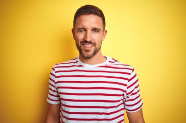 Sarı izole edilmiş arka plan üzerine sıradan kırmızı çizgili tişört giyen genç yakışıklı adam kameraya seksi bir ifadeyle bakıyor, neşeli ve mutlu bir yüzle..