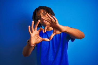 Rastalı tişörtlü Afro-Amerikan bir adam izole edilmiş mavi arka planda duruyor. Aşkla gülümsüyor, elleriyle kalp sembolü yapıyor. Romantik konsept.