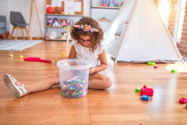 Gözlüklü ve tek boynuzlu at diademi takmış, yerde oturmuş anaokulundaki yapı taşlarıyla oynayan güzel bir çocuk.