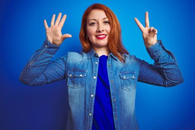 Genç, güzel, kızıl saçlı, kot gömlek giyen, mavi izole edilmiş arka plan üzerinde duran, yedi numaralı parmaklarıyla işaret eden, kendinden emin ve mutlu bir şekilde gülümseyen bir kadın..