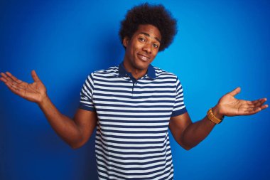 Afro saçlı, çizgili polo giyen, soyutlanmış mavi arka planda duran, kafası karışmış ve kolları ve elleri havada afro-amerikalı bir adam. Şüphe kavramı.