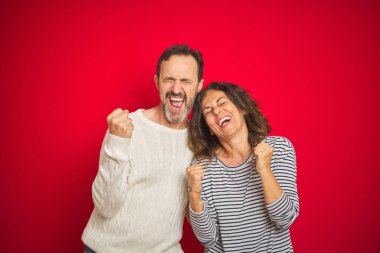 Güzel bir orta yaşlı çift, izole edilmiş kırmızı arka plan üzerine kışlık süveter giyip mutlu ve heyecanlı bir şekilde kollarını kaldırıp, gülümseyerek ve başarı çığlıkları atarak zafer jesti yapıyorlar. Kutlama konsepti.
