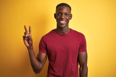 Kırmızı tişört giyen genç Afro-Amerikalı adam izole edilmiş sarı arka planda dikiliyor ve kameraya gülümsüyor zafer işareti yapıyor. İki numara..