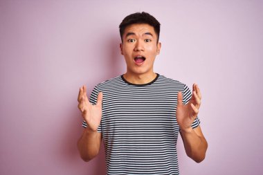 Asyalı genç bir Çinli, çizgili tişört giymiş, pembe arka planda durmuş çılgın ve hayret verici başarısını kutluyor, kollarını kaldırmış ve gözleri açık bir şekilde heyecanla haykırıyor. Kazanan konsept