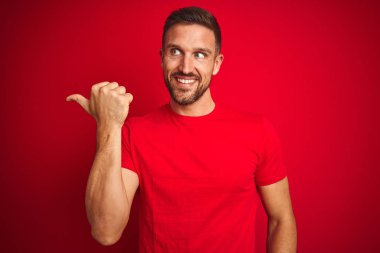 Kırmızı izole edilmiş arka planda rahat tişört giyen genç yakışıklı adam gülümsüyor ve başparmağıyla yan tarafa bakıyor..