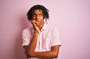 Zarif gömlekli Afro-Amerikan bir adam izole edilmiş pembe arka planda dikiliyor. Elleri ağızdan tırnak yerken gergin ve stresli görünüyor. Endişe sorunu.