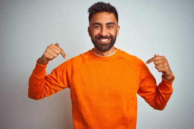 İzole edilmiş beyaz arka planda turuncu süveter giyen genç Hintli adam yüzünde bir gülümsemeyle gururla ve mutlu bir şekilde kendini işaret ediyor..