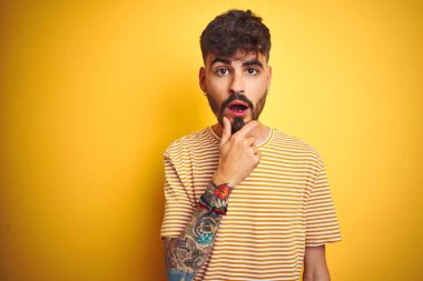 İzole edilmiş sarı arka planda duran çizgili tişörtlü genç adam, elleri çenesinde olan inançsızlığa, şaşkınlığa ve hayrete düşmüş ifadeye hayran görünüyor.