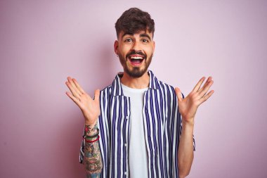 İzole edilmiş pembe arka plan üzerinde duran çizgili tişörtlü genç adam çılgın ve başarıyı kollarını kaldırıp heyecanla haykırarak kutluyor. Kazanan konsept