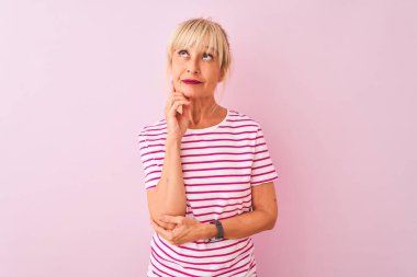 Orta yaşlı kadın çizgili tişört giyiyor pembe arka plan üzerinde duruyor, elleri çenesinde soru sormayı düşünüyor, düşünceli bir ifade. Düşünceli bir yüzle gülümsüyor. Şüphe kavramı.