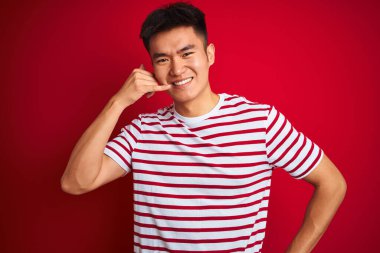 Asyalı genç bir Çinli, çizgili tişört giyip izole edilmiş kırmızı arka planda dikiliyor. El ve parmaklarıyla telefonla konuşuyor gibi gülümsüyor. İletişim kavramları.