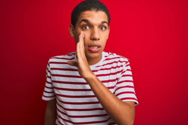 Young handsome arab man wearing striped t-shirt over isolated red background hand on mouth telling secret rumor, whispering malicious talk conversation