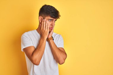 İzole edilmiş sarı arka planda duran beyaz t-shirt giyen genç Hintli adam. Yüzü, depresyonu ve üzüntüyü örten yorgun eller.
