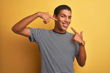 Genç yakışıklı Arap adam izole edilmiş sarı arka plan üzerinde donanma çizgili tişört giyiyor neşeli bir şekilde gülümsüyor ve parmaklarını ve ağzını işaret ediyor. Diş sağlığı konsepti.