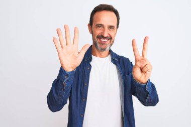 Mavi kot gömlek giyen orta yaşlı yakışıklı adam izole edilmiş beyaz arka plan üzerinde duruyor ve yedinci parmaklarıyla işaret ederken kendinden emin ve mutlu bir şekilde gülümsüyor..