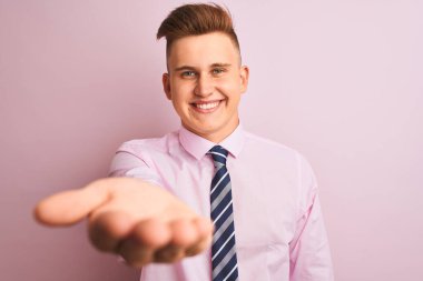 Genç yakışıklı işadamı gömlek ve kravat izole pembe arka plan üzerinde ayakta giyen neşeli sunan palmiye el yardım ve kabul veren gülümseyerek.