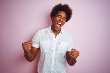 Afro saçlı, beyaz tişörtlü genç bir adam pembe arka plan üzerinde mutlu ve heyecanlı bir şekilde kollarını kaldırıp, gülümseyerek ve başarı için çığlık atarak zafer hareketi yapıyor. Kutlama konsepti.