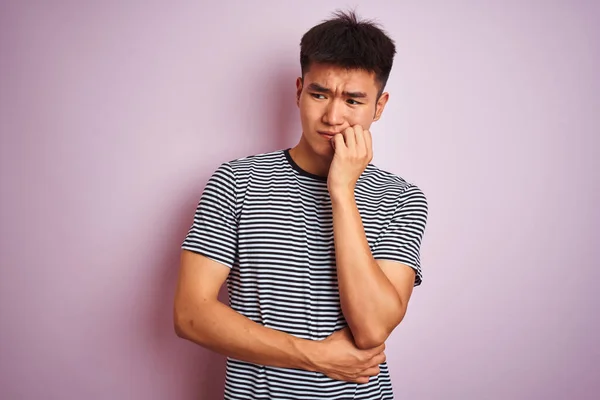 縞模様のTシャツを着た若いアジア系中国人男性が 口の中で爪をかむ上で手でストレスと緊張を見て隔離されたピンクの背景の上に立っている 不安の問題 — ストック写真