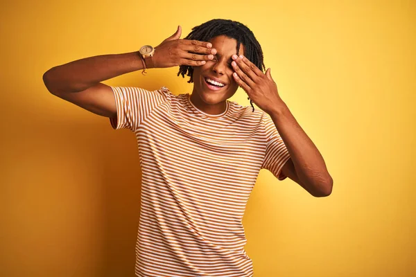 Afro man with dreadlocks wearing striped t-shirt standing over isolated yellow background covering eyes with hands smiling cheerful and funny. Blind concept.