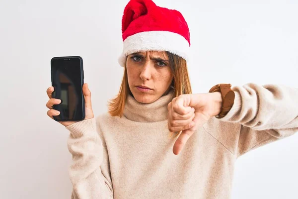 怒っている顔をしたスマートフォンの画面を示すクリスマスの帽子をかぶって美しい赤毛の女性 親指を下げて嫌いであることを示す負の記号 拒絶概念 — ストック写真