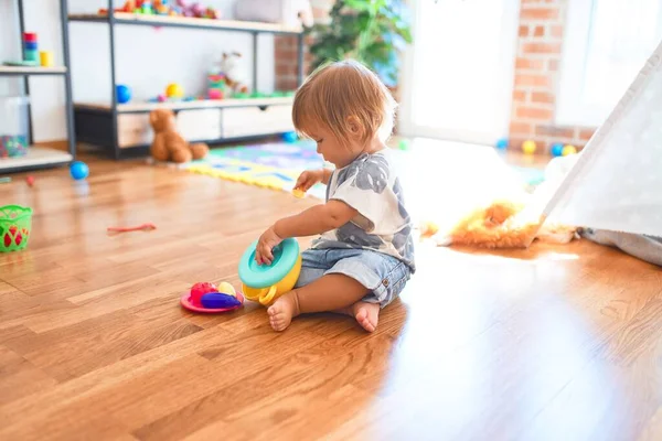 幼児は床に座ってプラスチック製のカトラリーや食べ物を使って食事をしています幼稚園のおもちゃの多くの周りに — ストック写真