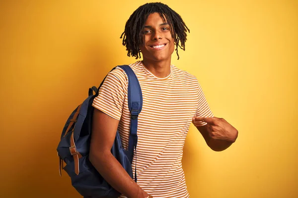 Afro american student man with dreadlocks wearing backpack over isolated yellow background with surprise face pointing finger to himself