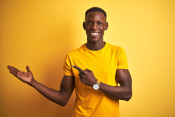 Young african american man wearing casual t-shirt standing over isolated yellow background amazed and smiling to the camera while presenting with hand and pointing with finger.