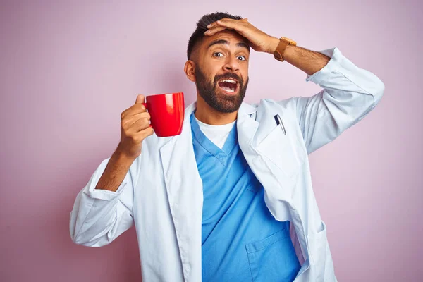 Young indian doctor man drinking cup of coffee standing over isolated pink background stressed with hand on head, shocked with shame and surprise face, angry and frustrated. Fear and upset for mistake.