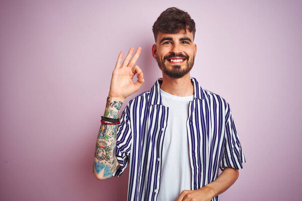 Young man with tattoo wearing striped shirt standing over isolated pink background smiling positive doing ok sign with hand and fingers. Successful expression.